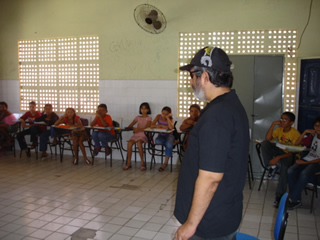 André está vestindo blusa preta, usando boné e óculos escuros. Ele está no centro da sala com as cadeiras posicionadas em forma de U, com as crianças de frente para ele. Os alunos esto divididos entre meninas e meninos numa faixa etária dos 9 aos 11
anos